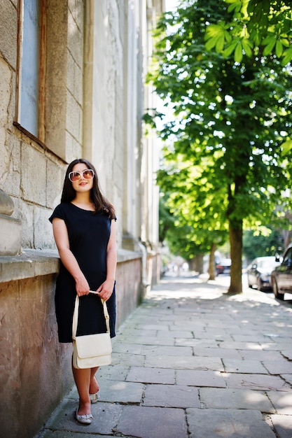 Brunette girl at black dress on sunglasses with handbag at hand posing at street of city