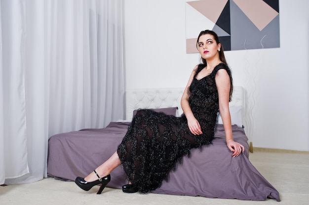 Brunette girl in black evening dress against white room with bed