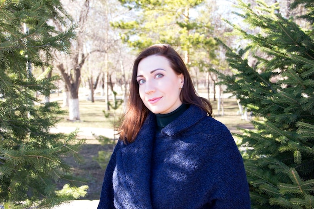 Brunette girl dressed in a blue coat in a park on a background of fir trees