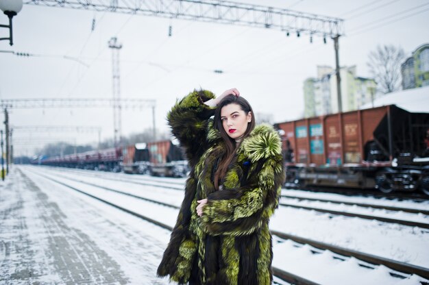 Brunette girl in green fur coat on the platform station at winter day.