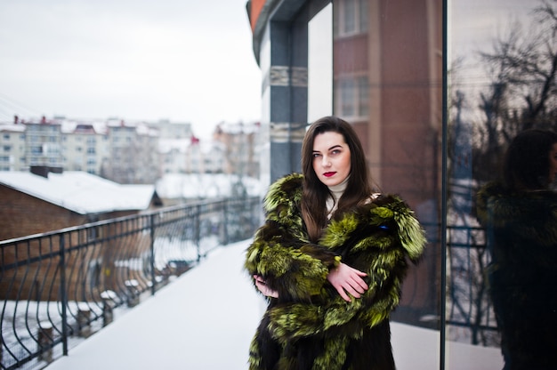 Brunette girl in green fur coat at street of city against house with large windows at winter.