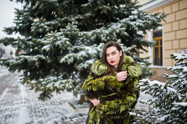 Brunette girl in green fur coat at winter day against snowy pine tree.