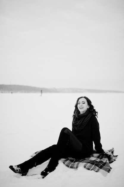 Brunette girl in green sweater and red scarf sitting on plaid outdoor frozen lake on evening winter day.