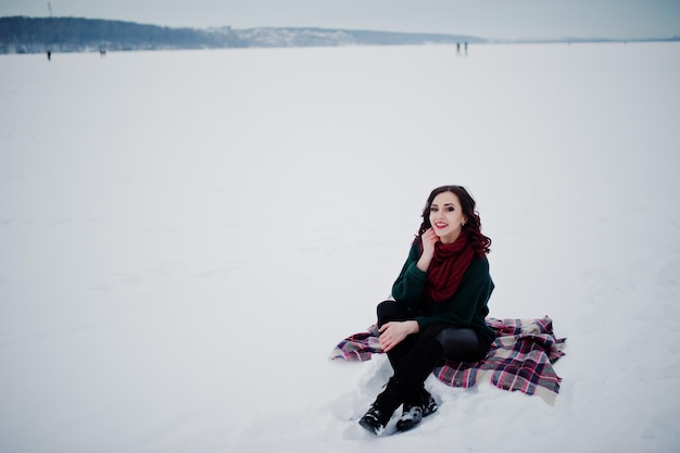 Brunette girl in green sweater and red scarf sitting on plaid outdoor frozen lake on evening winter day