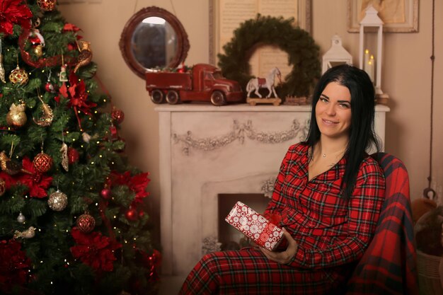 a brunette girl in pajamas is sitting on a chair by the fireplace with a gift box in her hands