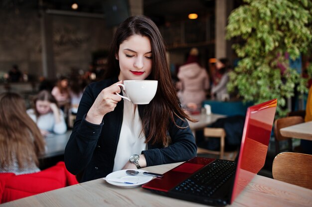 Brunette girl sitting on cafe with cup of cappuccino, working with red laptop.