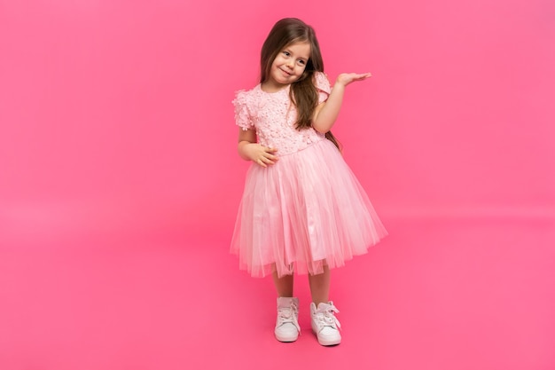 Brunette girl wearing pink dress amazed and smiling to the camera while presenting with hand isolated on pink.