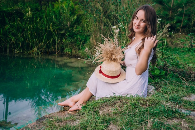 Brunette girl in a white dress and holding a straw hat near the lake with a bouquet of flowers.