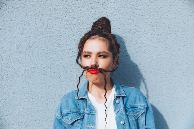 A brunette girl with dreadlocks and red lipstick in a denim suit is standing near a blue wall generation z