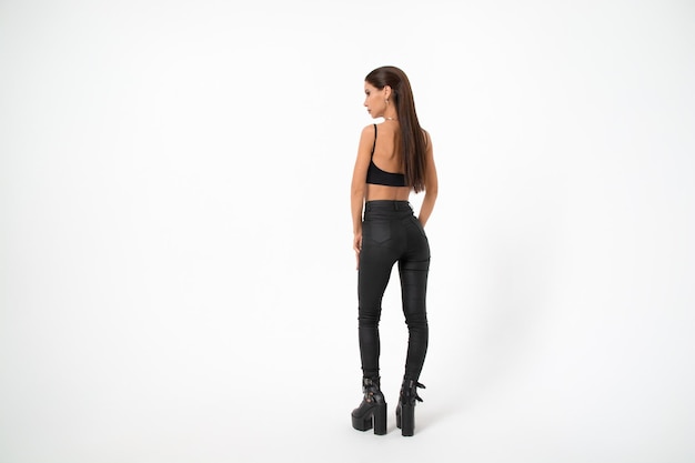 A brunette girl with long hair in black leather pants and boots poses on a white backgroundyouth fashion