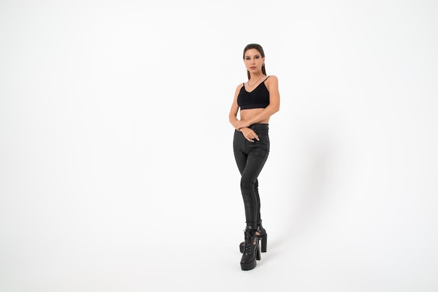 A brunette girl with long hair in black leather pants and boots poses on a white backgroundyouth fashion