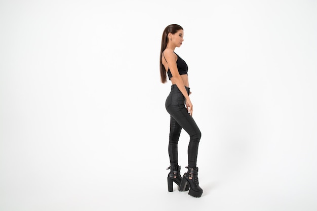 A brunette girl with long hair in black leather pants and boots poses on a white backgroundyouth fashion