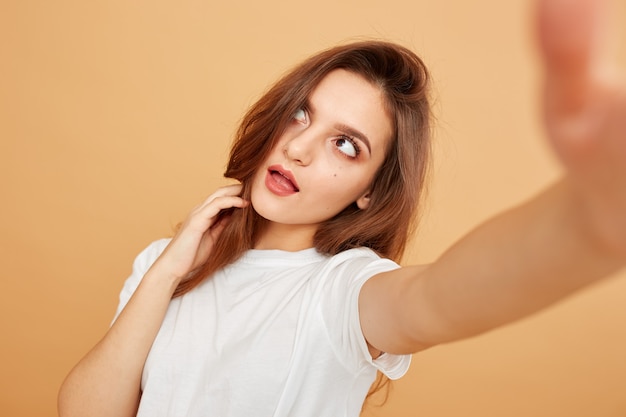 Brunette girl with long hair dressed in white t-shirt makes a selfie on the beige background in the studio .