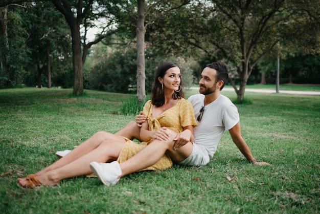 A brunette girl in a yellow dress is sitting between the legs of her boyfriend on the grass in old Spain town. A laughing couple of tourists on a date in the Valencian park.