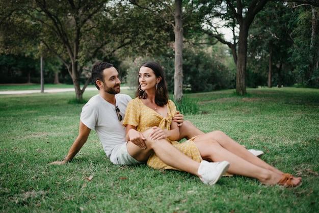 A brunette girl in a yellow dress is sitting between the legs of her boyfriend on the grass in old Spain town. A laughing couple of tourists on a date in the Valencian park.