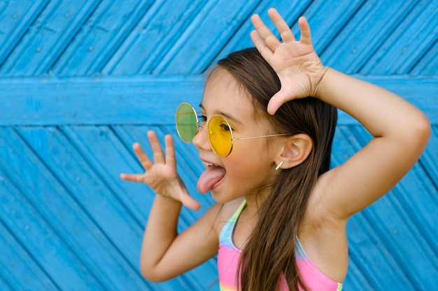 Brunette girl in yellow sunglasses stands against a blue wall and makes funny grimace showing tongue