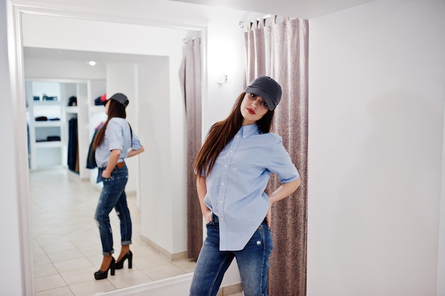 Brunette gorgeous girl in the clothing store boutique at casual clothes and cap against mirror