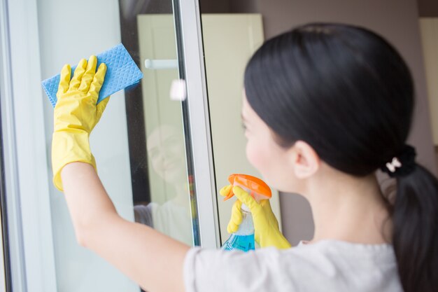 Brunette is cleaning the glass of window with blue rag and liquid spray. She does it careful.