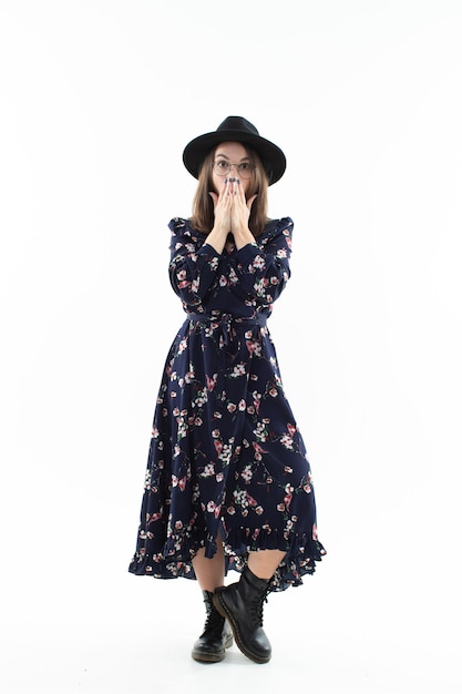 A brunette in a long floral dress with a hat poses for the camera in the studio