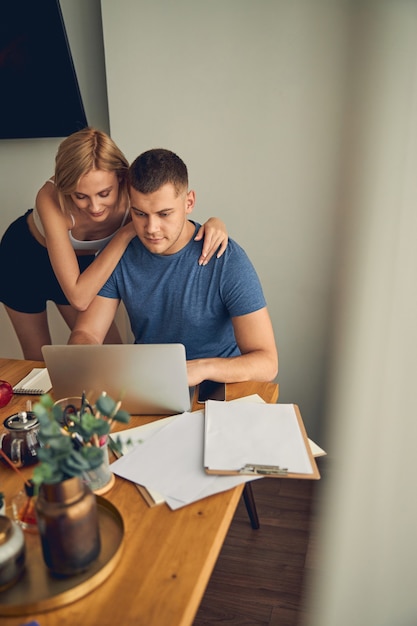 Brunette male working on laptop at home while cute young woman hugging and watching him