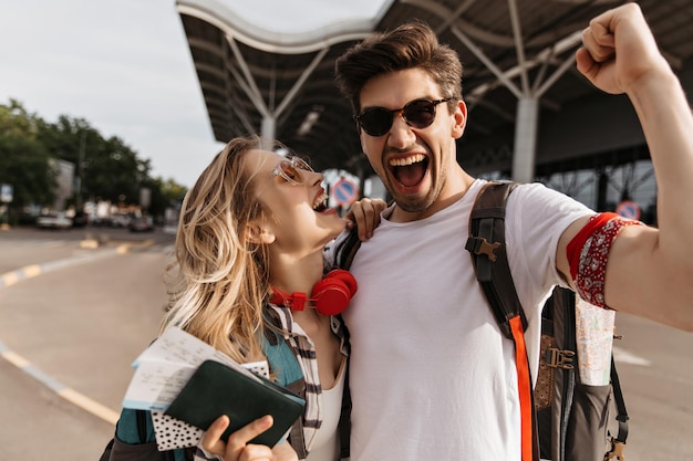 Brunette man happily screams and takes selfie with girlfriend near airport Cool blonde girl holds passport and laughs