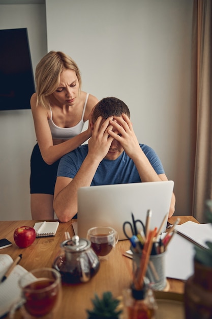 Brunette man sitting and feeling bad while blond slim woman in white top hugging him