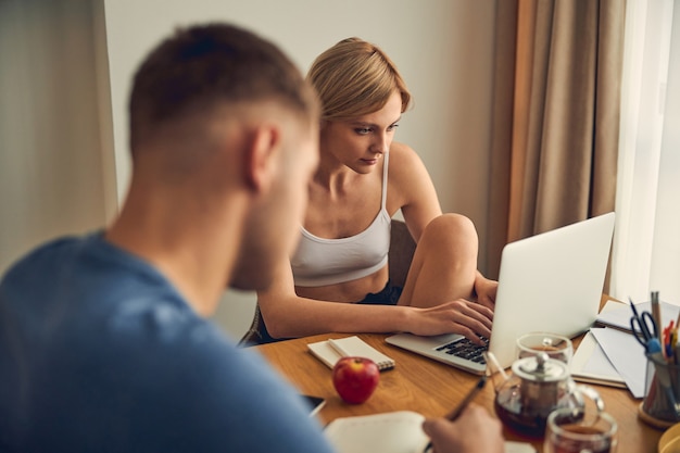 Brunette man sitting and writing while pretty woman spending time near device with notebook and pen