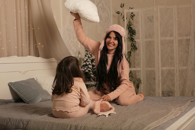 a brunette mother and daughter in pink pajamas are sitting on the bed and playing with a pillow