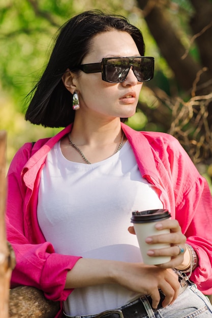 Brunette pretty woman in sunglasses holding coffee cup and walking in the park