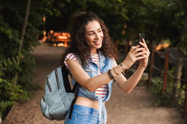 brunette pretty woman with backpack, smiling and photographing nature on smartphone while walking through green park