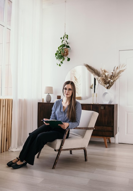 A brunette psychologist woman is sitting on a chair in the room and waiting for a patient's appointment. Professional psychologist in full growth