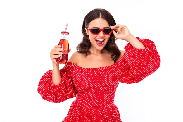 Brunette in a red dress with a bottle of fresh juice is dancing on a white background