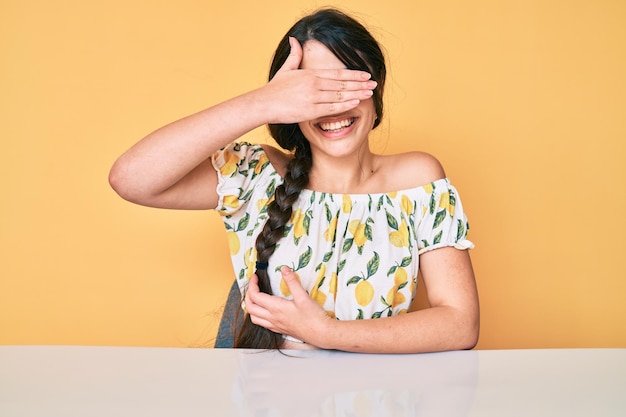 Brunette teenager girl wearing casual clothes sitting on the table smiling and laughing with hand on face covering eyes for surprise blind concept