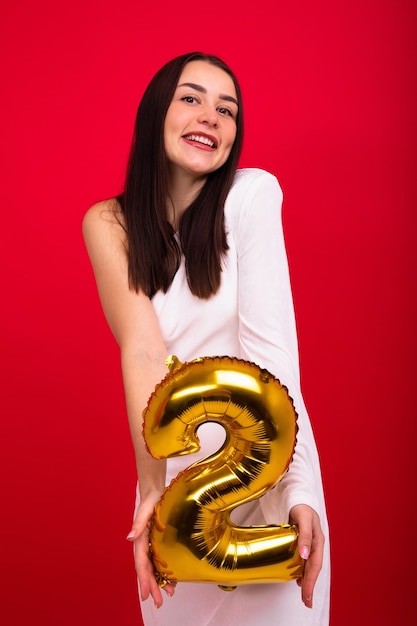 A brunette in a white dress holds a balloon in the shape of the number two on a red background