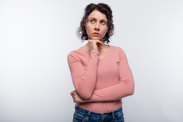 A brunette with curly hair in a pink blouse looks up thoughtfully