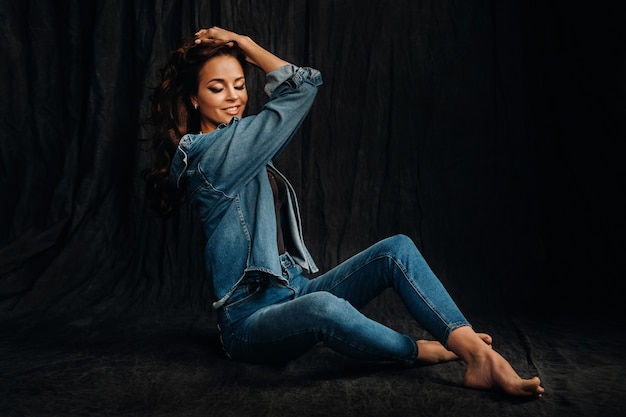 A brunette with long hair poses in a studio sitting on the floor on a black background.