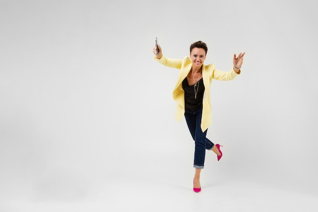 brunette with a short haircut in a cream-colored jacket and pink pumps pumps dancing with a phone in her hands on gray