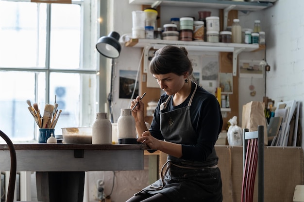 Brunette woman in black apron enjoys painting white handmade pottery sitting at table in workshop