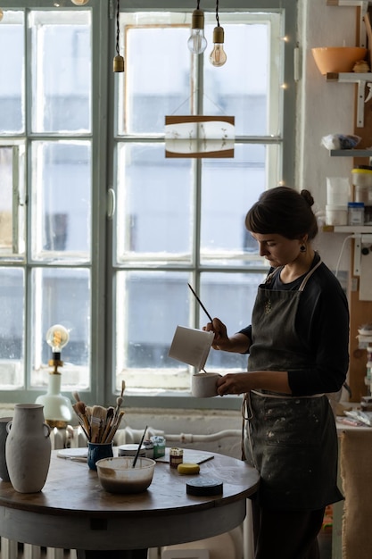 Brunette woman in black apron pours white paint into ceramic cup to work with pottery in workshop