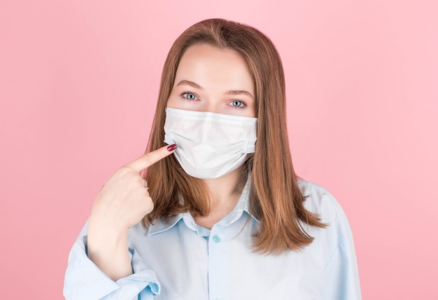 Brunette woman in a blue shirt wears a mask, shows a finger at herself on a pink