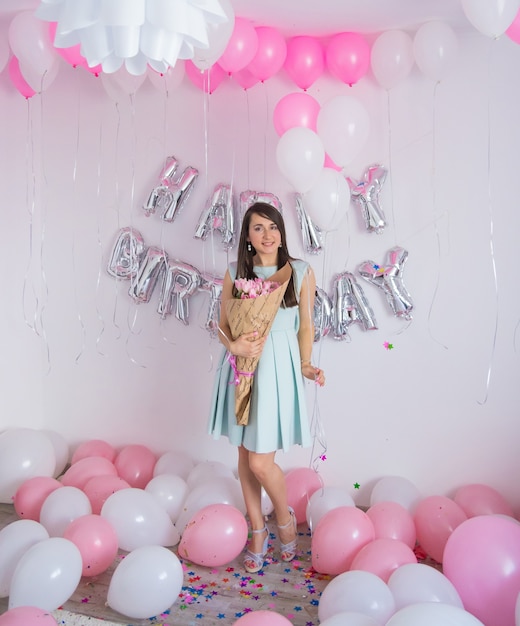 Brunette woman in mint dress holding bouquet of tulips. Birthday decorations with white and pink color balloons, bouquet of tulips and confetti for party on a white wall. Happy birthday anniversary.