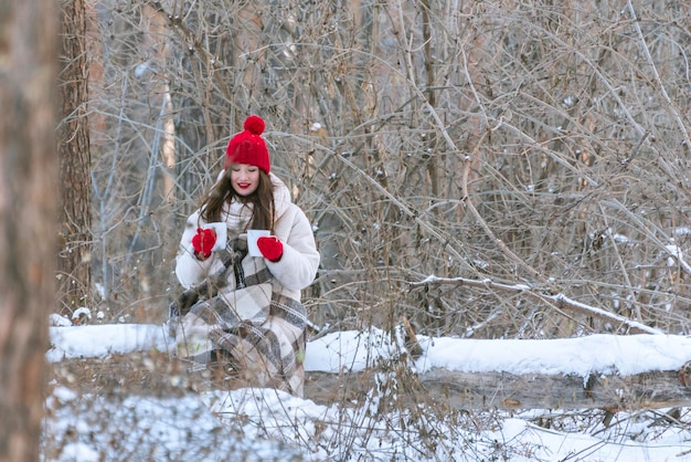 Brunette woman in red hat and mittens in winter forest with two mugs tea in anticipation of the beloved man