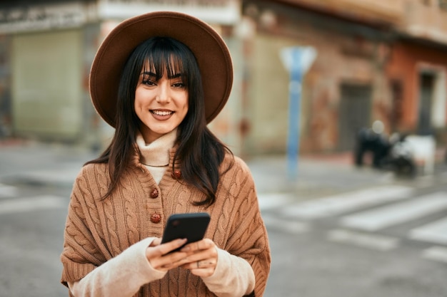 Brunette woman wearing winter hat smiling using smartphone outdoors at the city