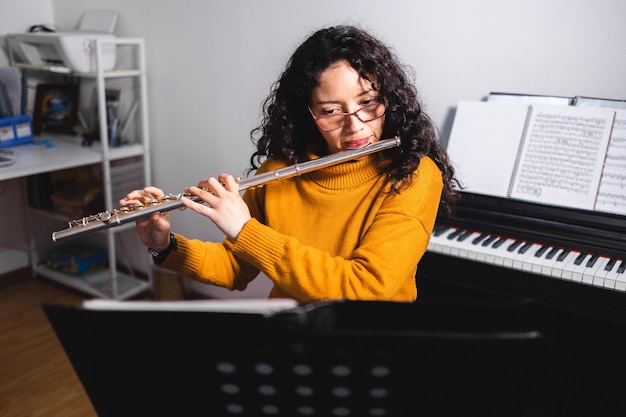 Brunette woman wearing a yellow sweater, and playing a transverse flute by reading sheet music.