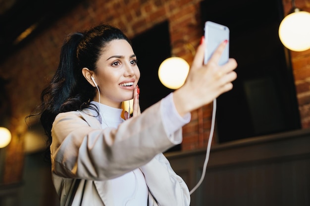 Brunette woman in white jacket having dark eyes with long eyelashes full lips and healthy skin having good mood while resting in cafe listening to different tracks with earphones and making selfie