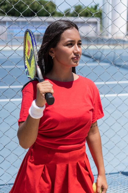 Brunette Woman with racket and ball standing near tennis court fence during break in training