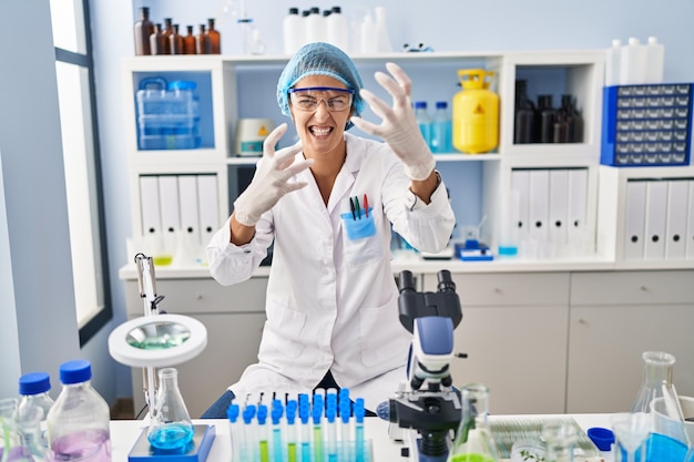 Brunette woman working at scientist laboratory shouting frustrated with rage, hands trying to strangle, yelling mad