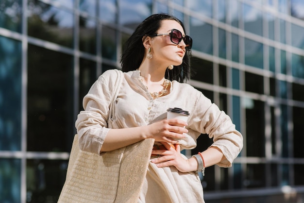 Brunette young woman with sunglasses and bag holding coffee walking in the city. Lifestyle portrait of woman