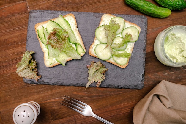 Bruschetta sandwich bread toast with soft green avocado cheese ricottafresh cucumbersbasil greenery on wooden cutting boardTasty healthy vegetarian foodItalian cuisine appetizertop view flat lay