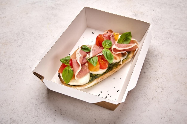 Bruschetta with mozzarella parma ham and pesto in white cardboard container on light background